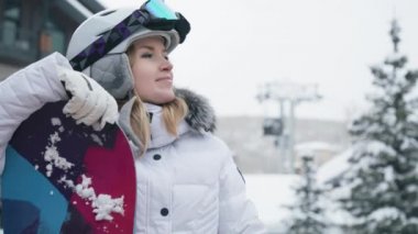 Slow motion 6K shot on RED camera happy woman with snowboard with ski lift on blurry background. Portrait of smiling cheerful woman in sports helmet and goggles enjoying winter day in ski resort USA