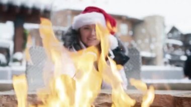 Beautiful young woman dressed warmly with fun red Santa Claus hat in winter ski resort village in the fresh frosty air roasting marshmallow in shape of snowman. Winter holidays slow motion background