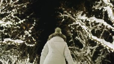 Woman in white winter coat walking by cinematic Aspen village. Slow motion back view woman enjoying snowy Christmas night with scenic illumination in city park on background. Holiday vacation 6K video