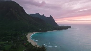 Sunset over ocean waves at sandy beach. Aerial rose golden sunset above mountain peaks silhouette. Tropical nature landscape. Cinematic drone shot paradise Kauai island coast, Hawaii USA travel 4K