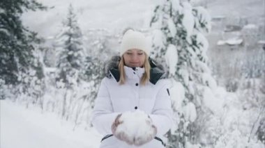 Slow motion young attractive woman looks into camera and throwing bunch of snow into air making fun out of being outdoors on snowy cold winter day. Beautiful woman laughing cheerfully having good time