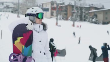 Snowboarder on background of ski slope at Snowmass ski resort. Slow motion close up portrait of athletic female tourist on snowy winter day with snowboarders shredding the slopes on blurred background