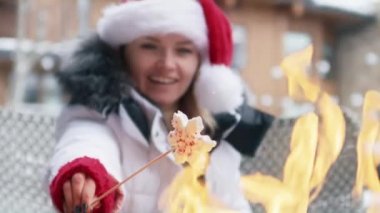Slow motion close up view smiling woman with marshmallow in snowflake shape with peppermint sprinkles. Woman in red Santa hat sitting by bonfire outdoors in modern ski resort Aspen during winter 6K