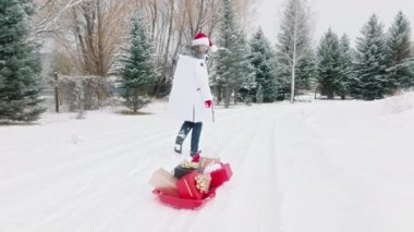 Happy woman in red Santa hat with large red sleighs full of gifts walking by scenic snowy spruce winter forest. Woman looks at gift boxes and hurry to congratulate people. Christmas New Year concept