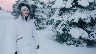 Slow motion young happy woman in warm white winter coat with fur hood walking in beautiful snowy spruce forest on colorful pink sunset sky background. Winter wonderland landscape on Christmas eve, RED