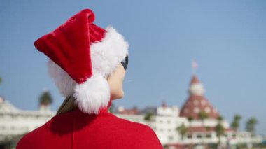 Happy New Year merry holiday concept. Back view of smiling woman in red sweater and Santa Christmas hat having fun outdoors on sunny winter day. Looking at copy space background isolated on blue sky