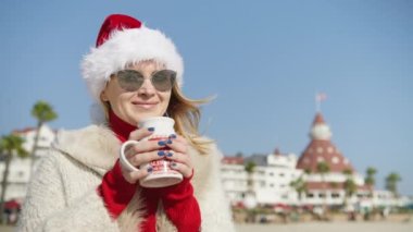 Portrait of satisfied woman drinking hot tea outdoors on sunny winter day. Attractive lady celebrating Christmas season enjoying cup of hot chocolate in slow motion. Smiling woman holding cup in hands