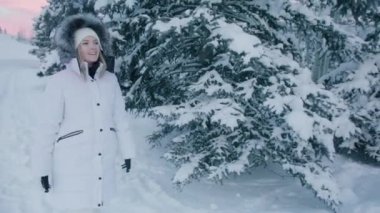 Portrait of young happy woman smiling with blonde hair under fur hood looking at pink sunset sky over snowy cold spruce forest wearing warm white winter puffer while walking in woodland, Slow Motion
