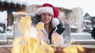 Attractive blonde woman sitting at outdoor bonfire in luxury ski resort hotel in Aspen village. Woman in red Santa Claus hat eating delicious marshmallows. Holidays concept slow motion shot on RED 6K