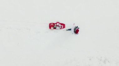 4K aerial top down winter background. Woman in Santa Claus costume walking by fresh white snow with red sled full of colorful gift boxes with beautiful golden and red bows on white Christmas eve day.