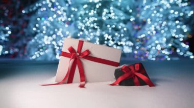 Magic night on Christmas eve with beautiful elegant gift boxes decorated with satin red ribbons. White and velvet black box on snow surface and glowing blue lights trees on blurry background, RED shot