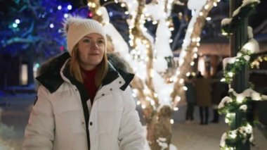 Portrait of nice smiling young woman enjoying cold Christmas season evening by walking in city park alley. Woman warming hands by breathing on cold fingers in fur mittens. Holiday vacation background