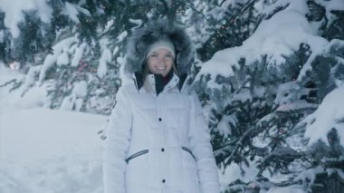 Portrait of attractive blue eyed smiling blonde confident pretty young woman in fur hood on white winter puffed coat standing under blue pine tree brunches covered by white snow of winter evening