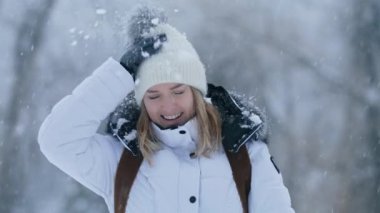 Happy positive blonde woman brushing off snow from beanie hat, laughing and having fun outdoors. Young woman enjoying freedom on winter vacation with snowy forest on blurred background, RED camera 6K