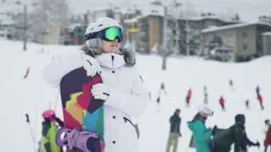 Happy smiling woman carrying her colorful snowboard after riding down the snowy mountain slope in Aspen village. Luxury hotels and ski resorts chalet buildings on blurry background, Snowmass village