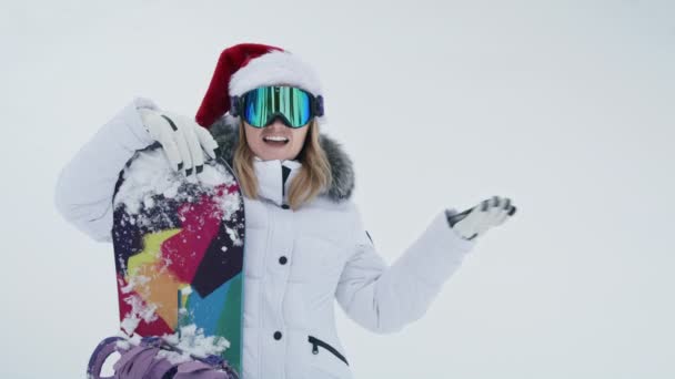 慢动作的30多岁女人戴着红色圣诞礼帽 指着雪白的纹理空间复制背景 模型与雪板有有趣的表达手势的手 新年假期概念6K — 图库视频影像