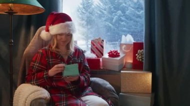 Kırmızı Noel Baba şapkalı güzel bir kadının portresi. Pencerede oturmuş Noel hediyeleri ve dışarıda şiddetli kar yağışı var. Mutlu kadın hediye için minnettar. Noel kutlaması 6K