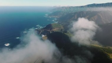 Derin mavi Pasifik okyanus manzaralı güzel bir dağ manzarası. Güneş ışınları beyaz bulutları aşıp kayalık kıyıya çıkıyor. Güneş ışığı dağ zirvelerine vurdu. İnsansız hava aracı bulutlarda uçuyor
