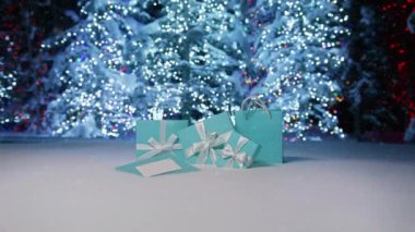 Parlak gümüş Noel ağaçlarına kar taneleri yavaşça düşüyor. Arka planda parlak ışıklar parlıyor. Sihirli gece, mutlu yeni yıl 2023 Zarif mavi hediye kutularının üzerinde beyaz kurdele görüntüleriyle kar yağışı