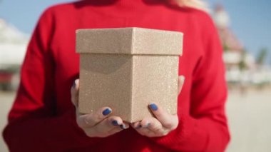 Holiday konsept çekimlerinin yavaş çekim görüntüsü. Kapalı kırmızı kazaklı kadın elinde altın gibi parlak bir kutuyla Noel hediyeleri ve güneşli kış gününde sürpriz hediyeler taşıyor.