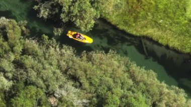 Parlak sarı pedallı teknede kürek çeken bir adamın yukarıdan aşağıya görüntüsü. Turist, yeşil ormandaki dere nehrinde hafta sonu su etkinliği sırasında eğleniyor. 4K erkek yüzme suyu taşımacılığı bisiklet ayak kontrolü