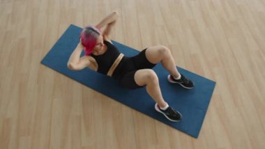 30 'larında etkin, sağlıklı bir kız paspasın üzerinde karın kaslarını çalıştırıyor. Pembe saçlı, beyaz bir kadın, yukarıdan görüldüğü gibi evde mekik çekiyor. Yüksek kalite 4k görüntü