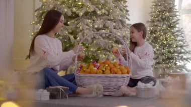 Kızlar Noel ağacının yanında otururken mandalina soyarlar. Neşeli kardeşler meyve yer ve gülerler. Yüksek kalite 4k görüntü