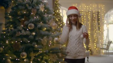 Beyaz bir kadın Noel ağacını süslerken telefonla konuşuyor. Mutlu kız geleneksel ağaca Xmas topu atıyor. Yüksek kalite 4k görüntü