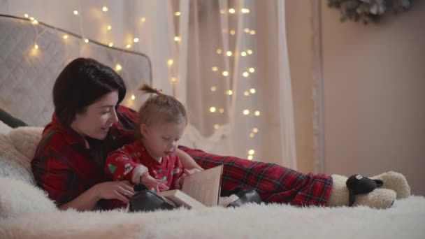 母と娘はクリスマスの衣装を着た本を読む 白人女性と彼女の子供は壁にクリスマスの照明とベッドの上に横たわっています 高品質4K映像 — ストック動画