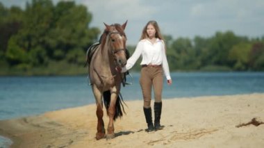 Beyaz bir kadın arka planda yemyeşil ormanlarla kumlu nehir kıyısında bir atla geziniyor. Güzel binici kadın binici büyüleyici bir hayvanla yürüyor. Yüksek kalite 4k görüntü