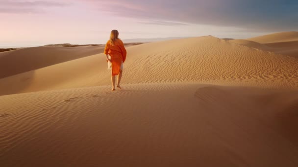 夕暮れ時に濃い灰色の雲とさざ波状の砂の砂漠の表面を歩く風のオレンジのドレスを振って裸足の女性旅行者 4K風光明媚な自然空中 女性のハイキング砂砂丘の映画的なスローモーション — ストック動画