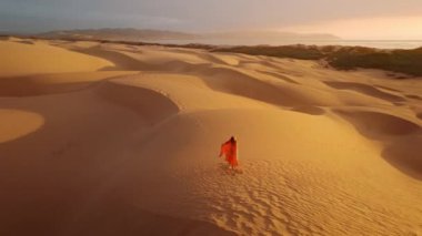 Kırmızı turuncu elbiseli kadın, elleri havada altın kum tepesinde dururken rüzgar elbisesini ve kızıl saçlarını üflüyor. Özgürlük, doğa konsepti. Kırmızıda sinemada altın gün batımı görüntüsü.