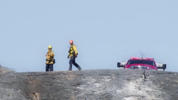 黄色の保護ヘルメットの男性消防士のシルエットは最近 暑い日に背景に乗って大きな赤い消防車で地上の丘に焼かれて歩く Redカメラで撮影された救助者の概念 — ストック動画