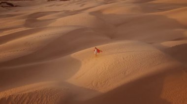 Uzun turuncu safari tarzı örtülü elbiseli kadın kum tepeciğinin üzerinde duruyor ve ellerini havaya kaldırıyor. Altın güneş silueti sinema arka planında parlıyor. Özgürlük mutluluğu ilham