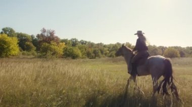 Kırsal alanda dört nala koşan şapkalı bir kadın. Atını eğiten sporcu kadın, binicilik kulübünün çevresindeki doğal manzaranın içinde. Yüksek kalite 4k görüntü