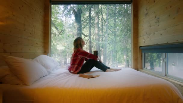 在山村里的小木屋里进行户外探险旅行的女人 森林景色迷人极了 穿着格子呢衬衫的旅行者坐在床上喝茶 望着窗户看书 幻想着生活 — 图库视频影像
