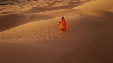 Gezgin, güzel doğa manzarası 4K ile destansı gün batımını izliyor. Turuncu elbiseli genç kadın altın gün batımında kum tepesinde koşuyor, kollarını havaya kaldırıyor, mutluluk, gençlik ve özgürlük kavramına.