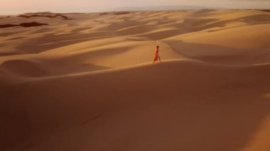 Macera, seyahat, mutluluk ve özgürlük kavramı. Altın günbatımında kum tepelerinin üstünde turuncu elbiseli bir kadının etrafında uçan yavaş çekim dronu. Güzel doğa manzaralı destansı günbatımında gezgin