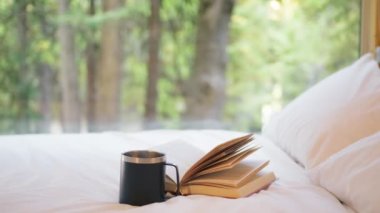 Siyah bardakta sıcak bir içecek ve boş beyaz yatak çarşafları üzerinde açık bir kitap. Sinema yeşil ormanı geniş bir pencereden görülüyor. Oda servisi, seyahat geçmişi, ekolojik otel, orman kulübesi.