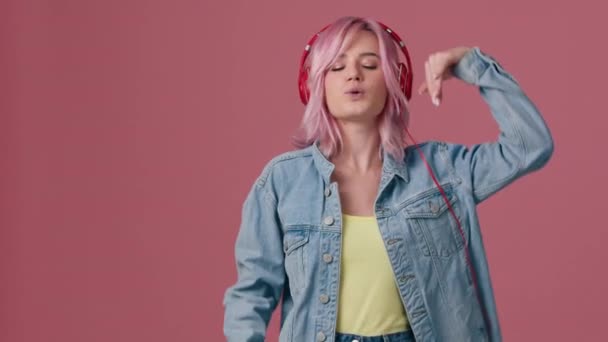 ピンクの背景にカラフルな髪型の若い女性 音楽を聴くと楽しくなる女性 20代モデルの笑顔 ピンクの髪が美しくなびく 女の子は音楽のリズムに移動します正の映像 — ストック動画