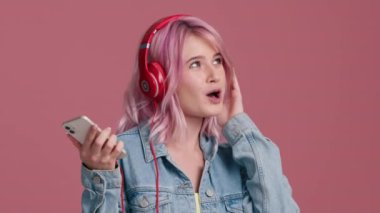 Kırmızı kulaklıklı, pembe saçlı, neşeli genç bayanın stüdyoda pembe izole edilmiş arka planda yüksek sesle dans edip eğlenmesi. İnsanlar, yaşam tarzı ve eğlence konsepti, modern teknolojiler