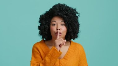 Sessiz bir jest. Mutlu Afro-Amerikan Kadın işaret parmağını dudaklara gösteriyor. Kıvırcık saçlı gülümseyen manken, mavi arkaplandaki kameraya bakarak sessizlik jesti yapar. İfade 4K 'ya söyleme