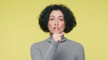 Çekici, olgun bir kadın, kameraya sarı arka planda sessizlik jesti yapıyor. İfade verme. Yetişkin bir kadın işaret parmağını dudaklarında tutar. Stüdyoda sessiz bir jest yapar.