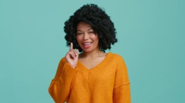 Koyu tenli neşeli bir kadın, mutlu bir şekilde seni kameraya ve olumlu göz kırpmalarına işaret ediyor. Mavi stüdyo arka planında izole edilmiş turuncu süveter giymiş, gülümseyen bir Afro-Amerikan modeli.