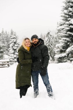 Çekici bir adam ve kadın soğuk ormanda sarılıyorlar. Kabarık ceketli yetişkin çift yürürken iyi eğlenceler. Kış zamanı romantik bir tarih. Kış aşk hikayesi.