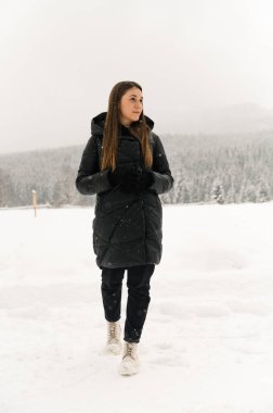 Kar yağışı altında yürüyen güzel bir kız. Siyah kabarık ceketli genç bir kadın. Kışın ormanda poz veriyor. Açık havada esmer bir bayanın portresi. Kış konsepti