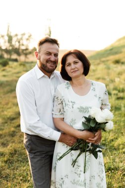 Orta yaşlı bir adam, elinde kocasının çiçekleri olan Avrupalı görünümlü bir kadına sarılıyor. Gün batımında aşık bir çiftin portresi.