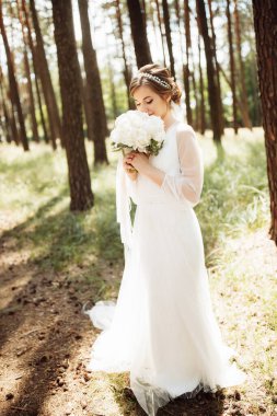 Ormanda zarif bir gelin duruyor ve elinde şakayıkla güzel bir düğün buketi tutuyor.