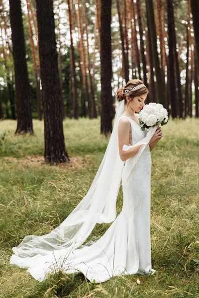 Ormanda zarif bir gelin duruyor ve elinde şakayıkla güzel bir düğün buketi tutuyor.