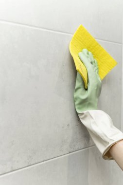 Duvardaki fayansları yıkıyorum. Lastik eldivenli bir kadın eli ve duvardaki fayansları sarı bir bez parçasıyla yıkıyor.